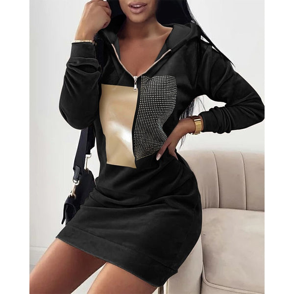 Vrouw in zwarte hoodie-jurk met beige en zwarte vierkante print, zittend op beige bank. Modieuze dameskleding, casual stijl.