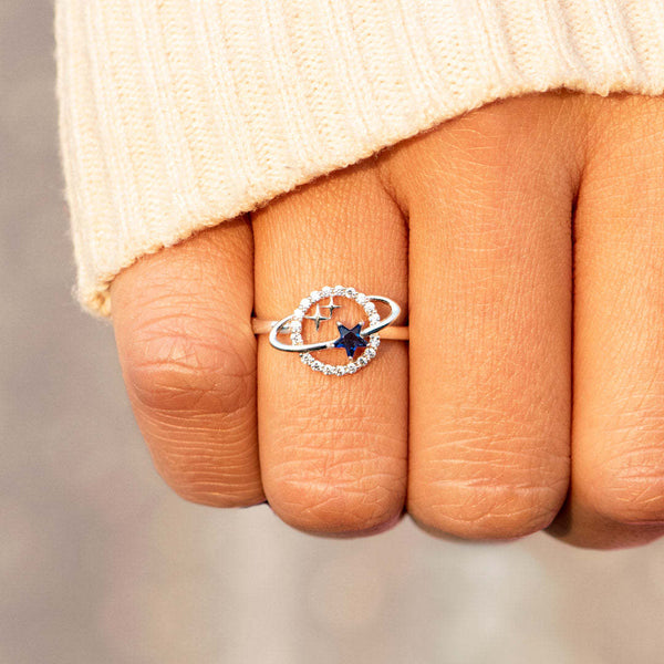 Zilveren ring met ster en cirkelvormig ontwerp, ingelegd met blauwe en heldere stenen, gedragen op een hand met beige trui. Elegante sieraden.
