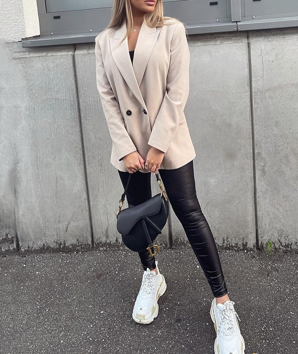 Vrouw in beige blazer en zwarte leren legging met witte sneakers en zwarte schoudertas, modieuze straatstijl, stedelijke achtergrond.