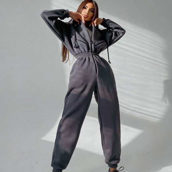 Vrouw in grijze joggingpak poseert tegen witte achtergrond, modieuze sportkleding, comfortabele loungewear, trendy damesmode, casual outfit.