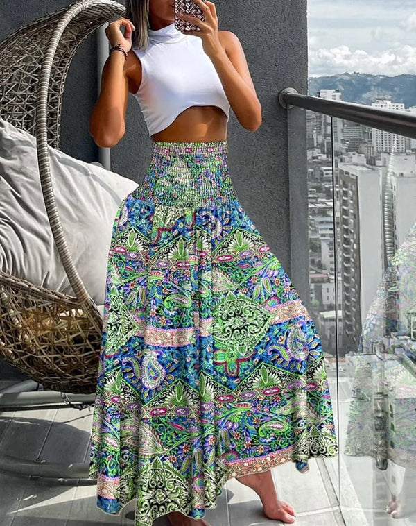 Vrouw in witte crop top en kleurrijke boho maxi-rok met paisley-patroon, staand op balkon met stadzicht. Zomermode, bohemian stijl.