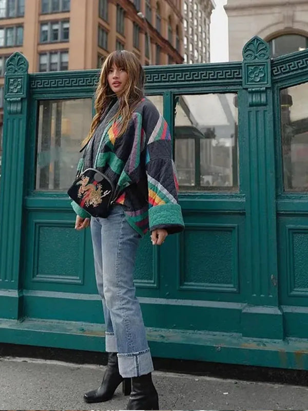 Vrouw in kleurrijke poncho en jeans voor metro-ingang in stedelijke omgeving, draagt zwarte laarzen en tas met draakontwerp. Trendy herfstmode.