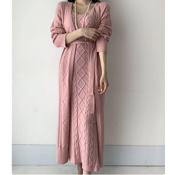 Vrouw in roze gebreide maxi-jurk met kabelpatroon, lange mouwen en ceintuur, tegen een witte achtergrond. Elegante damesmode, herfstkleding.