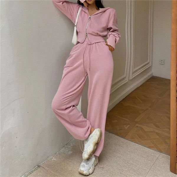 Vrouw in roze joggingpak leunt tegen muur, draagt witte sneakers. Comfortabele loungewear, casual mode, trendy dameskleding.