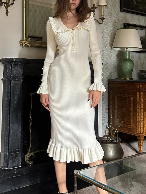 Witte geribbelde midi-jurk met lange mouwen en ruches, gedragen door een vrouw in een klassieke woonkamer. Elegante damesmode, herfstkleding.