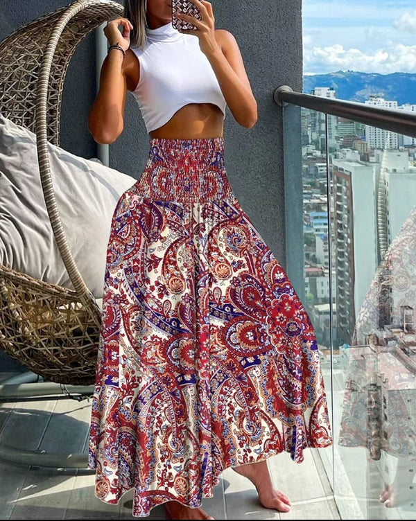 Vrouw in witte crop top en kleurrijke bohemian maxi-rok met paisley-patroon, staand op balkon met stadzicht. Zomermode, boho-stijl, trendy outfit.