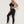 Vrouw in zwarte sportkleding, bestaande uit een sportbeha en legging, poseert tegen een witte achtergrond. Fitnessmode, actieve levensstijl.