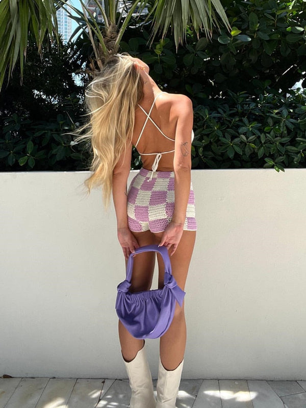 Vrouw in geblokte paarse en witte jurk met paarse tas en witte laarzen, poseert buiten voor groene planten. Zomermode, trendy outfit.