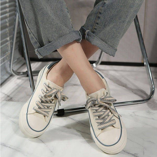 Witte canvas sneakers met dikke zolen en grijze veters, gedragen door persoon in opgerolde jeans, zittend op marmeren vloer. Trendy casual schoenen.