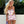 Blonde vrouw in geblokte paarse en witte gehaakte top en shorts, poseert buiten voor groene planten. Zomermode, trendy outfit, strandkleding.