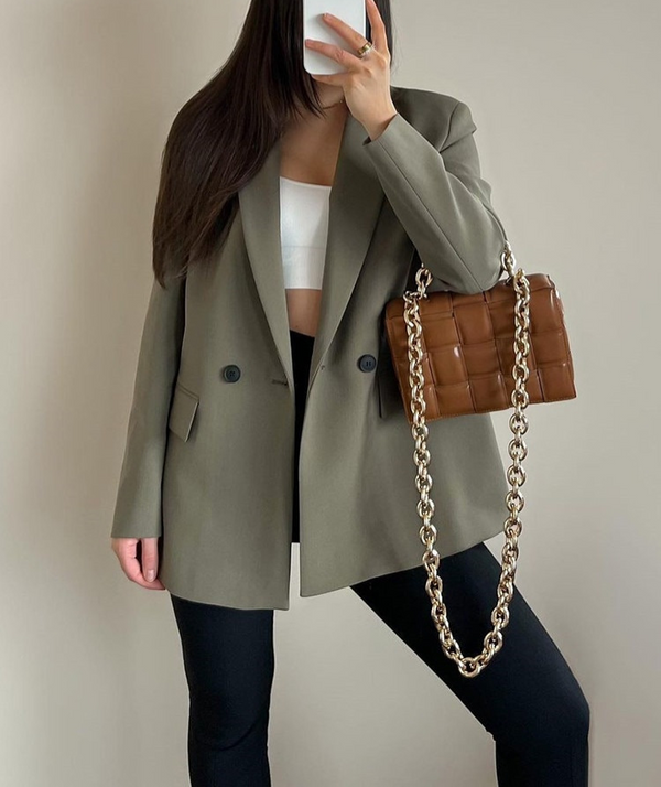 Vrouw in oversized groene blazer met witte crop top, zwarte broek en bruine gewatteerde tas met gouden ketting. Modieuze dameskleding.