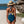 Vrouw in blauwe badpak en strohoed op zandstrand met rotsen en zee op achtergrond, zomermode, strandkleding, vakantie aan zee.