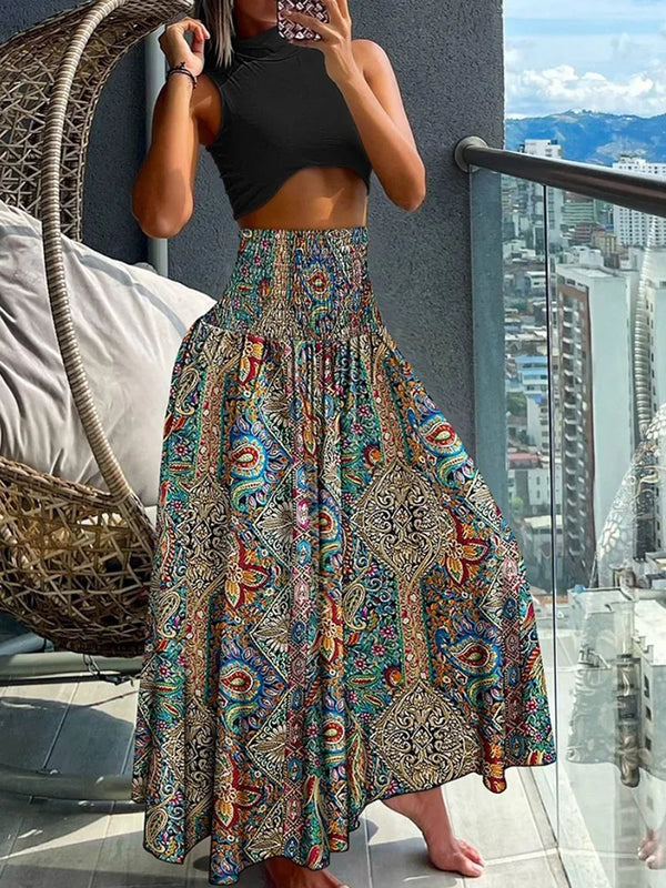 Vrouw in kleurrijke boho-maxirok en zwart crop top op balkon met stadzicht. Bohemian mode, zomeroutfit, stedelijke achtergrond.