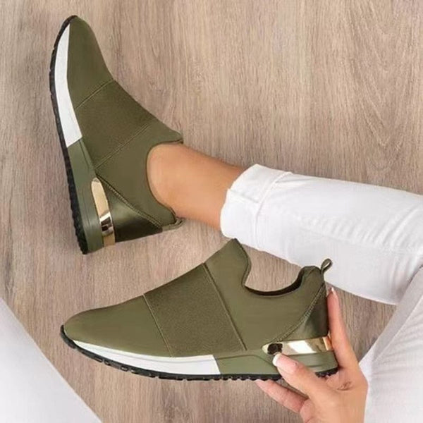 Olijfgroene slip-on sneakers met witte zool en gouden details, gedragen door persoon in witte broek op houten vloer. Comfortabele damesmode.