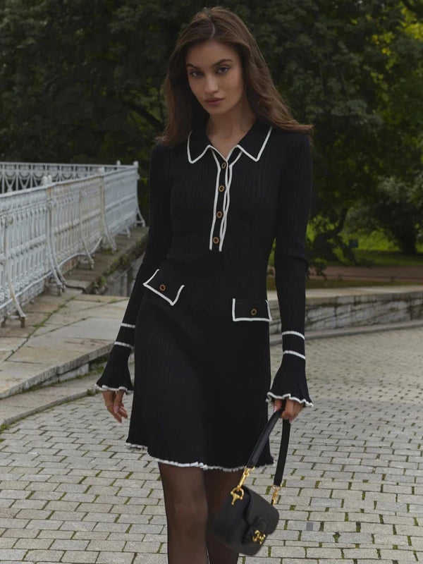 Vrouw in elegante zwarte jurk met witte accenten, knopen en lange mouwen, draagt een zwarte handtas, wandelend op een stenen pad in een park.