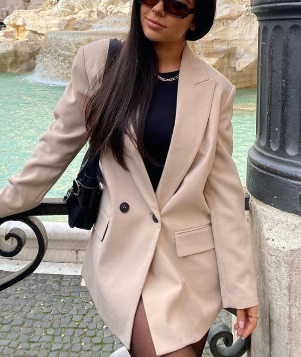 Vrouw in beige blazer bij fontein, draagt zonnebril en zwarte tas. Stijlvolle mode, stedelijke achtergrond, trendy outfit, casual chic look.