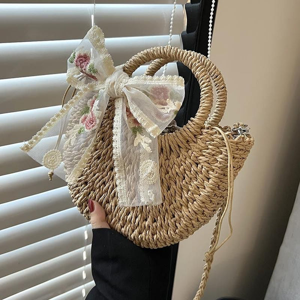 Rieten handtas met ronde vorm, versierd met een grote kanten strik met bloemenmotief. Perfect voor zomerse outfits en stranduitjes.