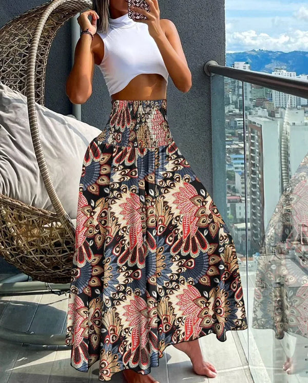 Vrouw in witte crop top en kleurrijke bohemian maxi-rok op balkon met stadzicht. Zomerse mode, boho-stijl, trendy dameskleding.
