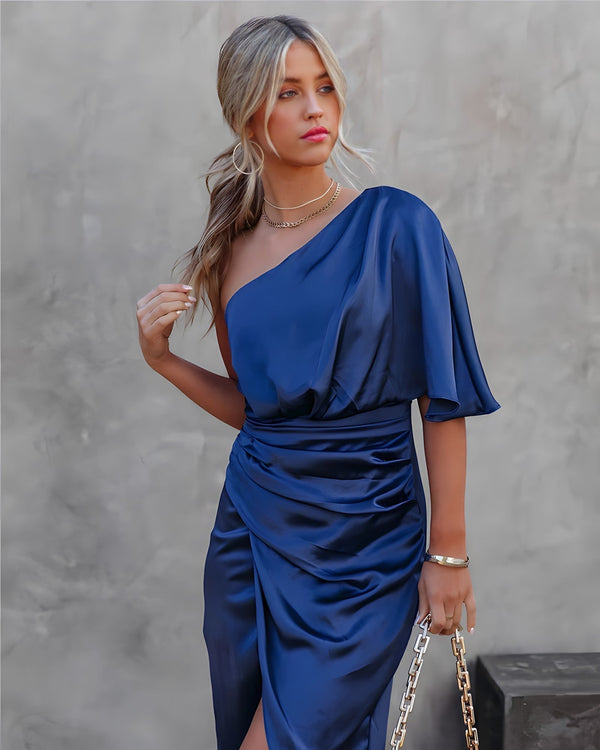 Vrouw in elegante blauwe satijnen asymmetrische jurk met gouden ketting en armband, tegen een neutrale achtergrond. Perfect voor avondmode en galakleding.