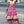 Vrouw in witte top en lange roze bohemian rok met etnisch patroon, draagt een strooien tas en sandalen, loopt op stoep in stedelijke omgeving.