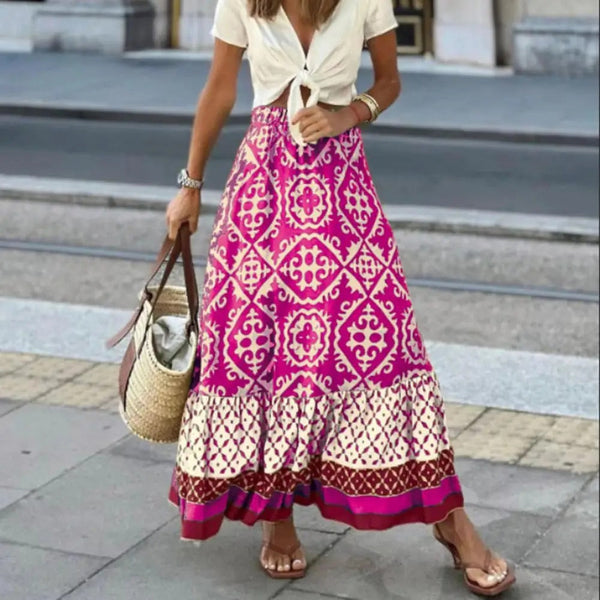 Vrouw in witte top en lange roze bohemian rok met etnisch patroon, draagt een strooien tas en sandalen, loopt op stoep in stedelijke omgeving.