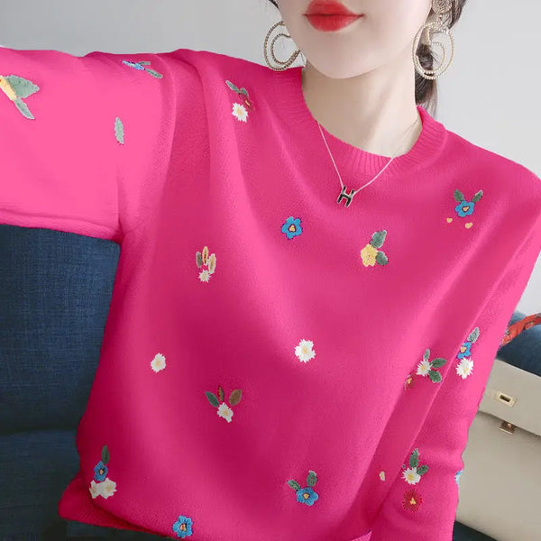 Vrouw in felroze trui met kleurrijke bloemenborduursels, zittend op een blauwe bank. Trendy damesmode, casual outfit, herfstkleding.