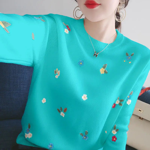 Vrouw draagt turquoise trui met kleurrijke bloemenborduursels, gouden oorbellen en ketting. Modieuze dameskleding, trendy herfststijl.
