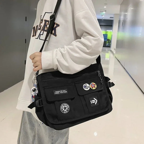 Persoon met witte trui draagt een zwarte schoudertas met patches en pins in een moderne gang. Casual mode, streetwear, trendy accessoires.