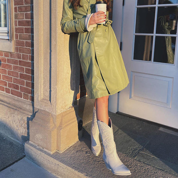 Vrouw in groene trenchcoat met witte cowboylaarzen en koffiebeker, leunend tegen bakstenen muur bij deur. Stijlvolle herfstmode, buiten.