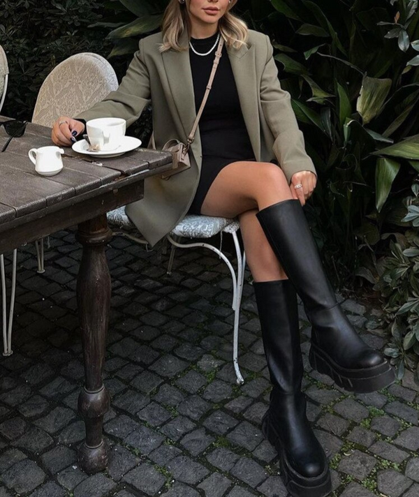 Vrouw in groene jas en zwarte laarzen zit aan houten tafel met koffiekopjes, omgeven door groene planten. Stijlvolle herfstmode, buitenomgeving.