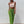Vrouw in beige asymmetrische top en groene gehaakte broek met uitgesneden details, draagt paarse zonnebril en witte laarzen, tegen witte achtergrond.