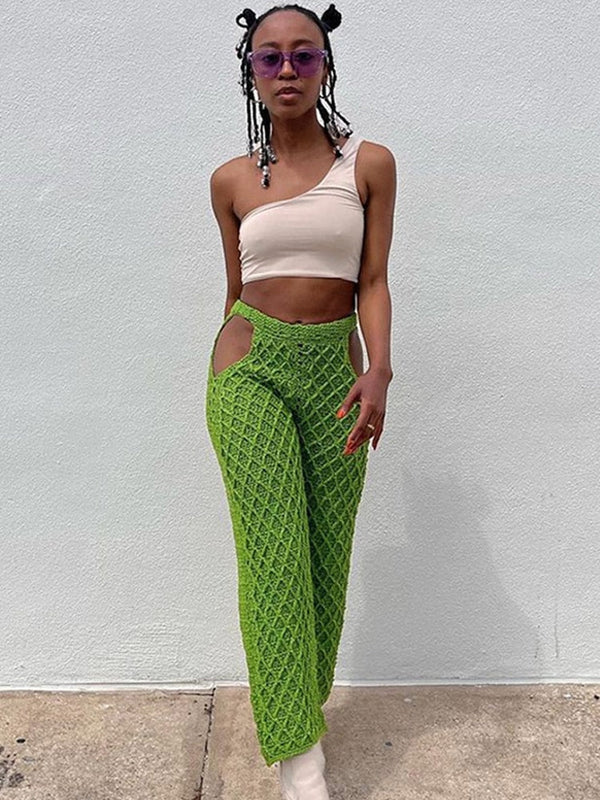 Vrouw in beige asymmetrische top en groene gehaakte broek met uitgesneden details, draagt paarse zonnebril en witte laarzen, tegen witte achtergrond.