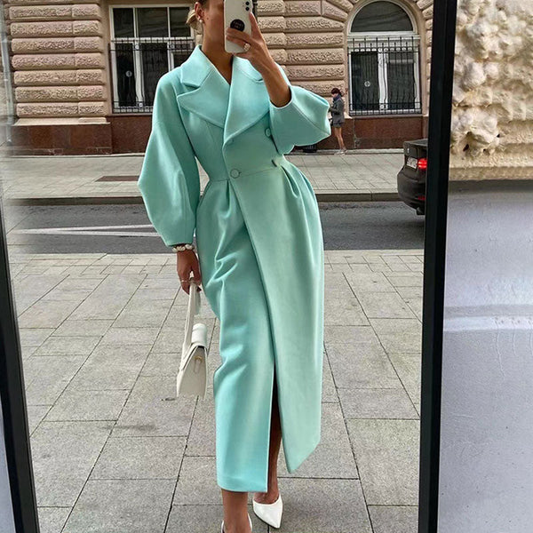 Vrouw in mintgroene lange jas met ceintuur, witte handtas en hakken, poseert voor spiegel op straat. Stijlvolle mode, elegante dameskleding.