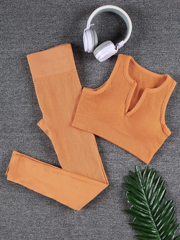 Oranje naadloze sportset met legging en crop top op grijze achtergrond, naast witte koptelefoon en groene palmblad. Perfect voor yoga en fitness.