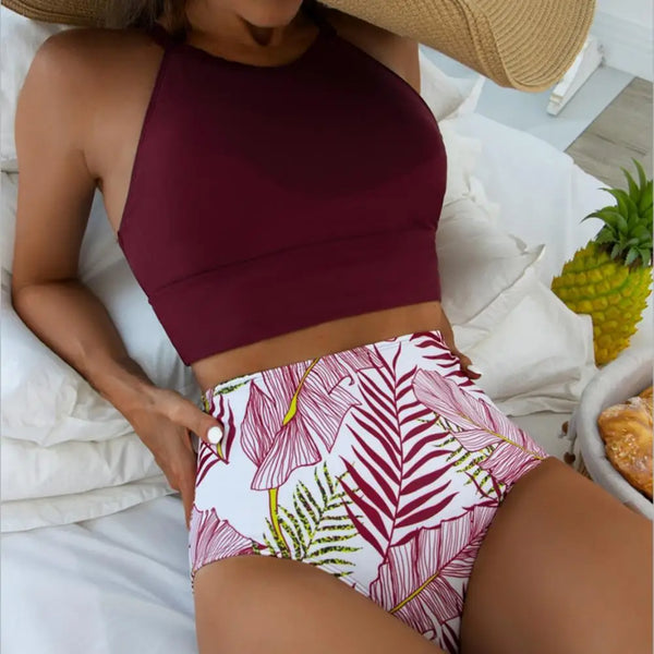 Vrouw in bordeauxrode crop top en tropische hoge taille bikini met bladerenprint, liggend op wit beddengoed naast ananas en hoed.