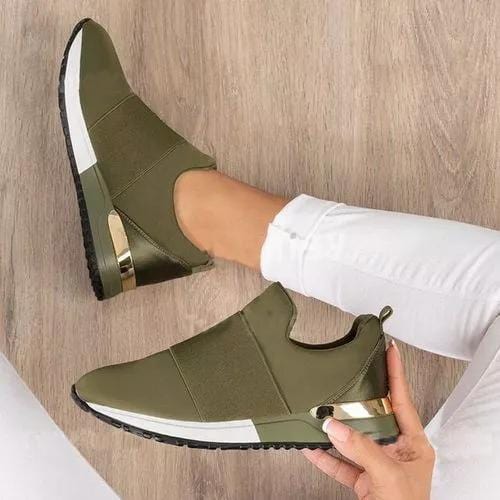 Olijfgroene sneakers met elastische band en gouden details, gedragen door persoon in witte broek op houten vloer. Comfortabele damesmode schoenen.