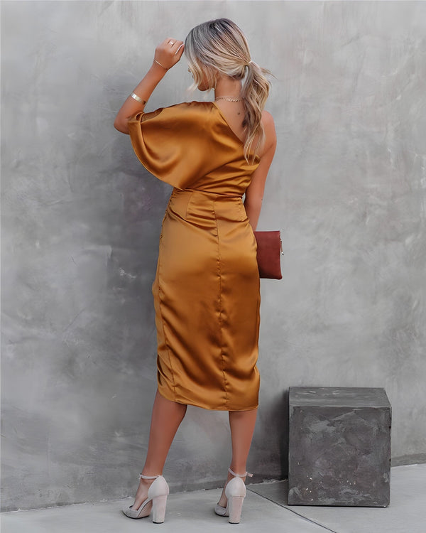 Vrouw in elegante, goudkleurige satijnen jurk met asymmetrische schouder, poseert tegen betonnen muur. Stijlvolle mode, avondkleding, chique uitstraling.