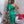Vrouw in groene asymmetrische jurk voor versierde kerstboom, omringd door cadeaus en kerstdecoraties, in moderne woonkamer.