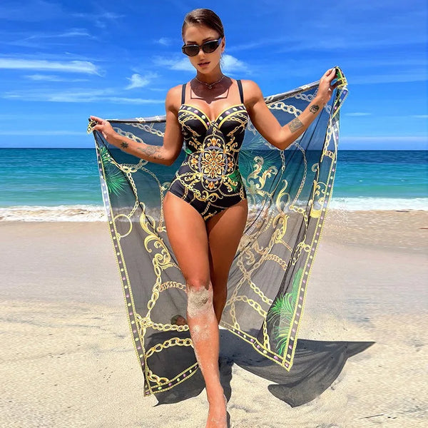Vrouw in elegante zwarte badpak met gouden kettingprint en doorschijnende omslagdoek, poseert op tropisch strand met blauwe lucht en zee.