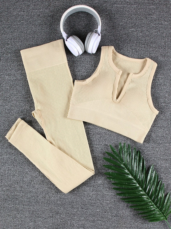 Beige sportset dames met legging en crop top, geribbelde stof, naast witte koptelefoon en groene palmblad, op grijze achtergrond.