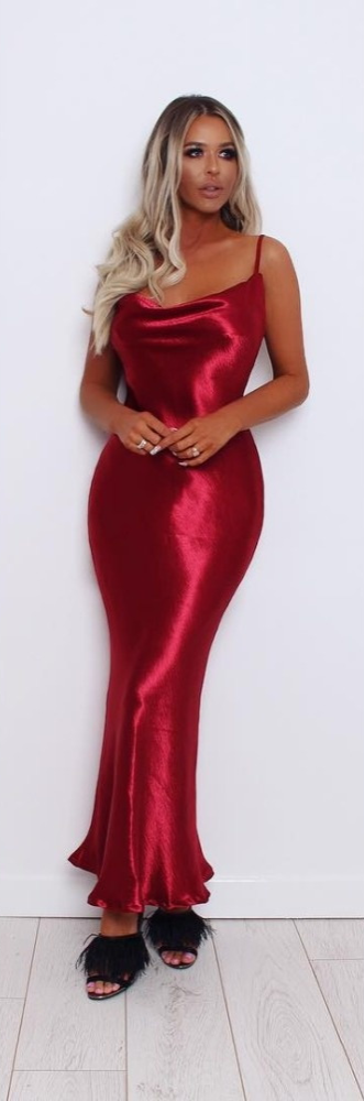 Vrouw in rode satijnen jurk met spaghettibandjes, staand tegen witte muur. Elegante avondjurk, perfect voor feestelijke gelegenheden.