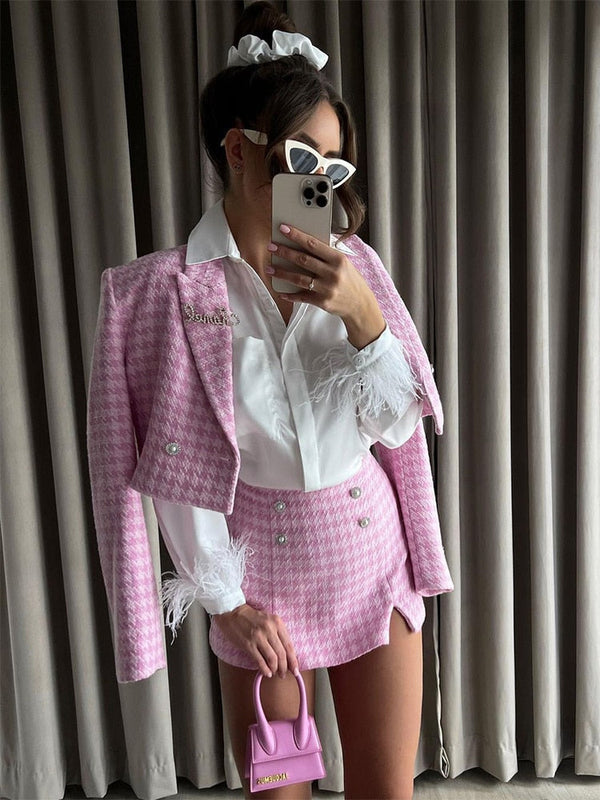 Vrouw in roze pied-de-poule pak met witte blouse en veren, houdt roze mini-handtas en smartphone vast, draagt zonnebril en scrunchie.