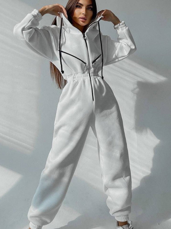 Vrouw in witte sportieve jumpsuit met capuchon, zwarte trekkoorden en elastische taille, poseert tegen een neutrale achtergrond.
