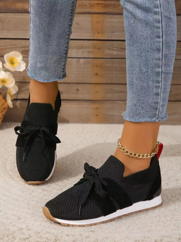 Zwarte dames sneakers met strik, gebreide stof, witte zool en bruine details, gedragen met blauwe jeans. Comfortabele en modieuze schoenen.