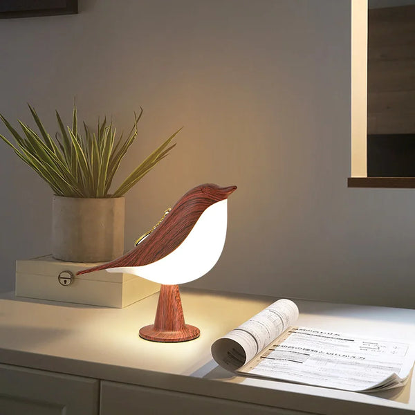 Moderne tafellamp in de vorm van een vogel, gemaakt van hout en glas, geplaatst op een witte tafel naast een plant en opgerolde krant.