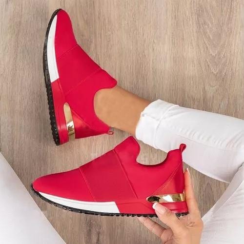 Rode slip-on sneakers met witte zool en gouden details, gedragen door persoon in witte broek op houten vloer. Comfortabele en modieuze damesschoenen.
