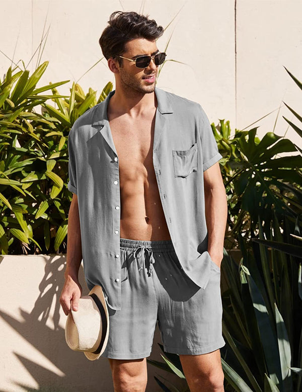 Man in grijs zomershort en overhemd, zonnebril, houdt hoed vast, omringd door groene planten. Casual herenkleding, zomermode, strandstijl.