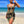 Vrouw in elegante strandjurk met kettingprint, zwarte en gouden details, poseert op tropisch strand met blauwe zee en heldere lucht.