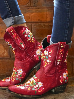 Rode cowboylaarzen met kleurrijke bloemenprint, gedragen door persoon in spijkerbroek, tegen bakstenen muur. Trendy damesmode, westernstijl.