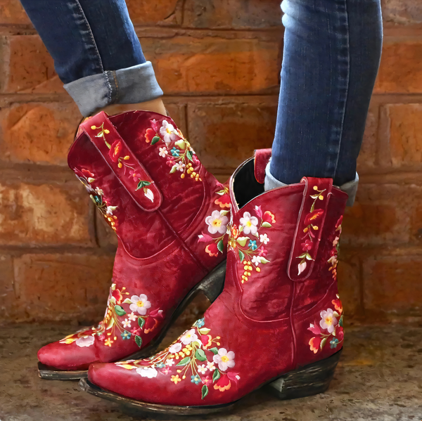 Rode cowboylaarzen met kleurrijke bloemenprint, gedragen door persoon in spijkerbroek, tegen bakstenen muur. Trendy damesmode, westernstijl.